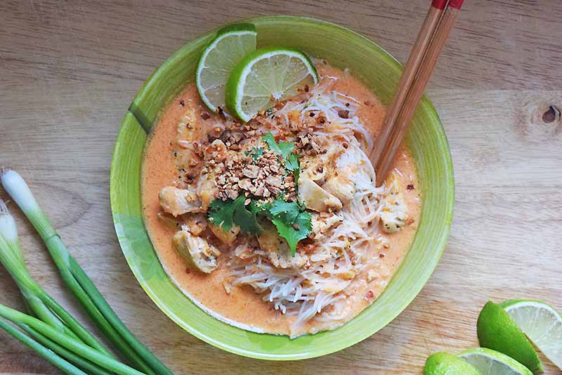 A top down picture of a green bowl with chicken and noodles in a creamy red curry sauce, topped with chopped nuts, cilantro, and slices of lime. To the right of the bowl are two wooden chopsticks, and lime slices. To the right of the bowl are some bunching onions. The background is a wooden surface.