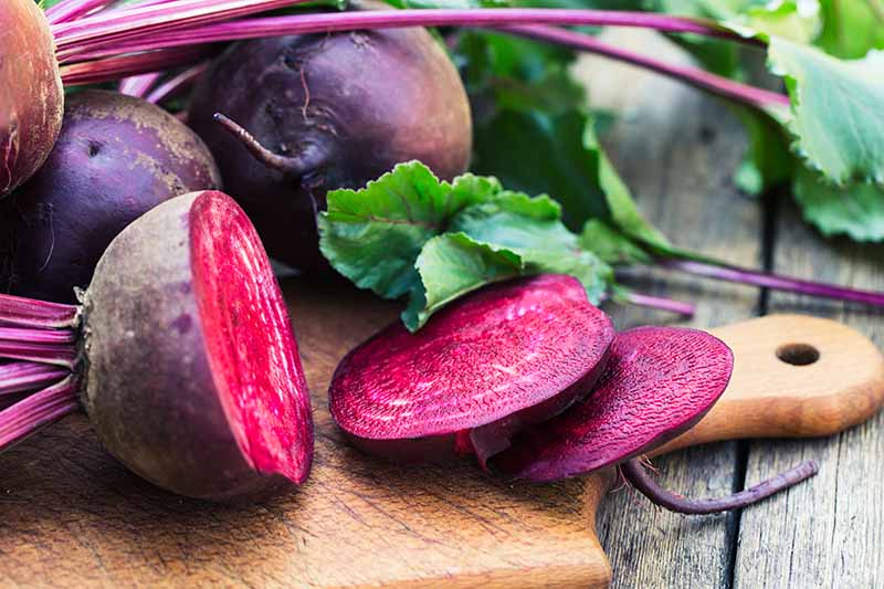 A wooden chopping board on a wooden surface, with a sliced beetroot and two whole ones behind. Some purple stems and foliage around the vegetables.