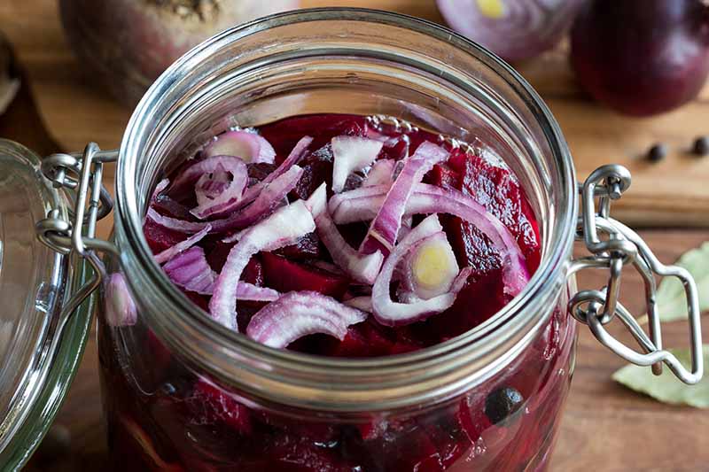 A close up looking into a jar with chopped beets in liquid with sliced red onion on the top. In the background is a wooden surface with onions fading to soft focus.