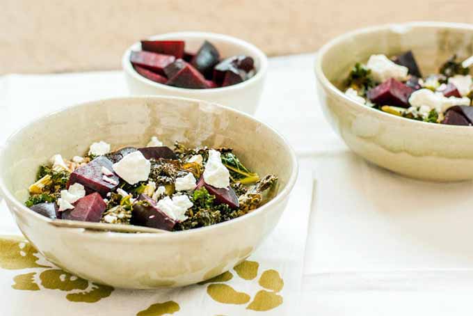 Two bowls of coconut, ginger, kale, goats cheese and beets, on a white and gold fabric placemat. Behind is a small white bowl with beetroot slices. The background is a white table cloth.