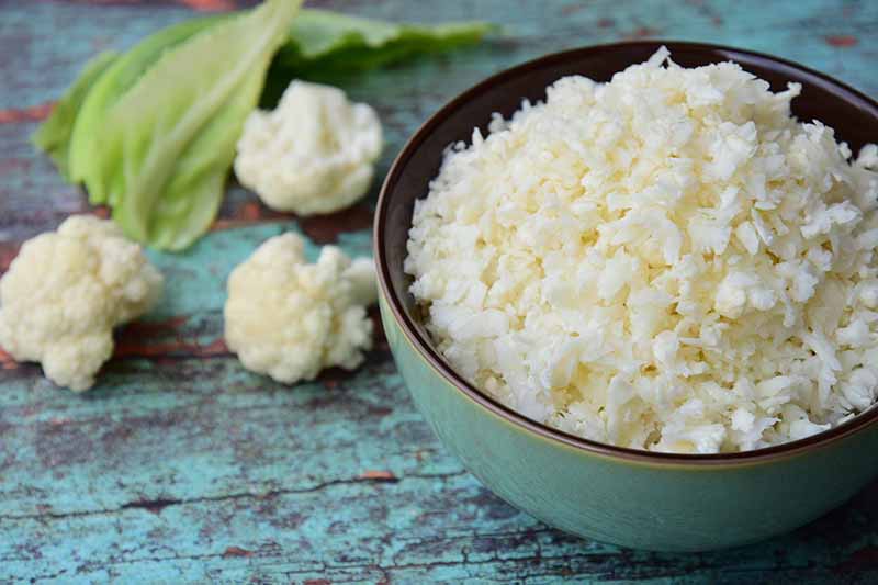 A close up of a blue ceramic bowl containing cauliflower rice, set on a rustic blue wooden surface, with a scattering of florets to the left, in soft focus in the background.