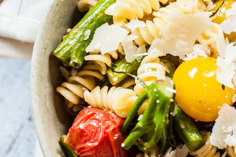A close up of a rustic bowl of pasta salad with grilled tomatoes, broccolini, and parmesan cheese, set on a blue surface.