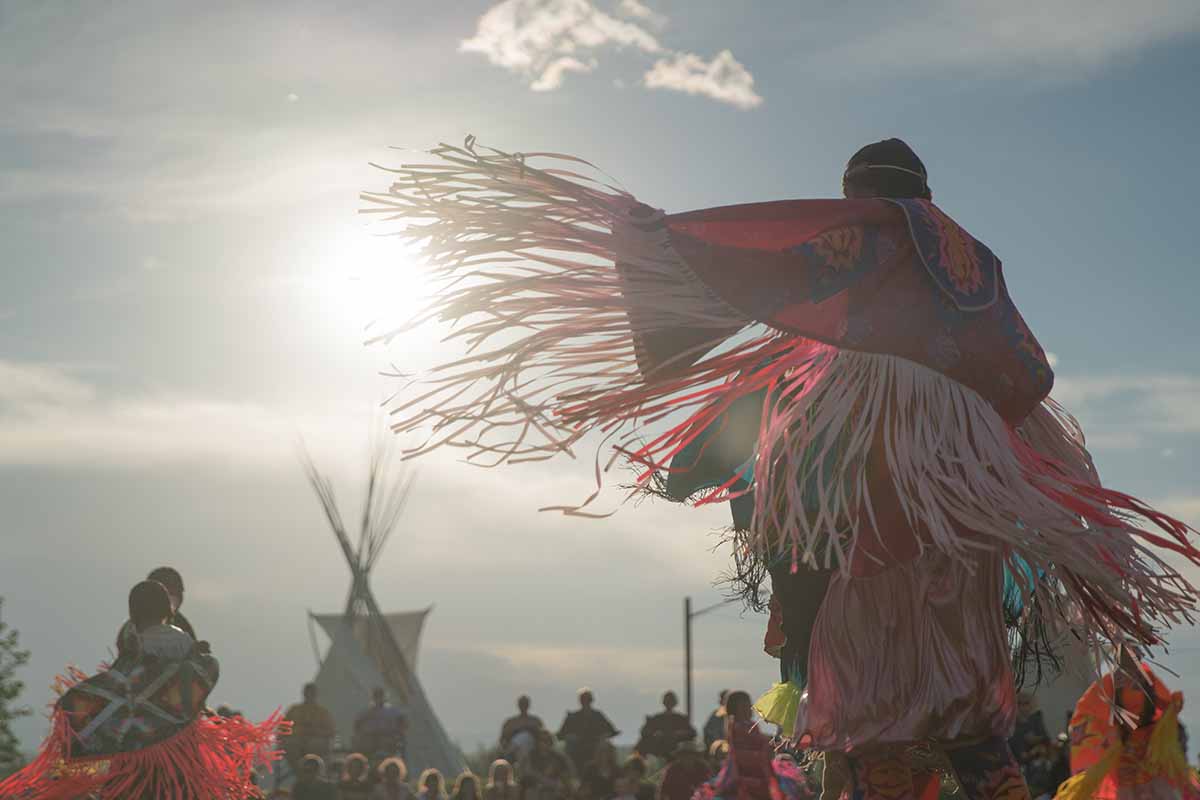 A horizontal shot of a an indigenous person dressed in Native American attire performing a rain dance. In the background is a traditional Native American teepee.