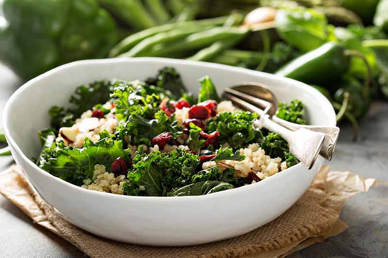 A white bowl containing a beautiful salad of raw Brassica oleracea and quinoa, with a spoon and fork in the side. Under the bowl is a piece of hessian and brown paper, on a gray surface. In the background, various green vegetables in soft focus.