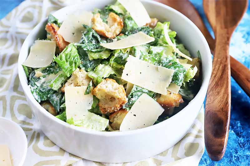 A close up horizontal image of a white bowl with a freshly made Caesar salad with croutons and shaved cheese.