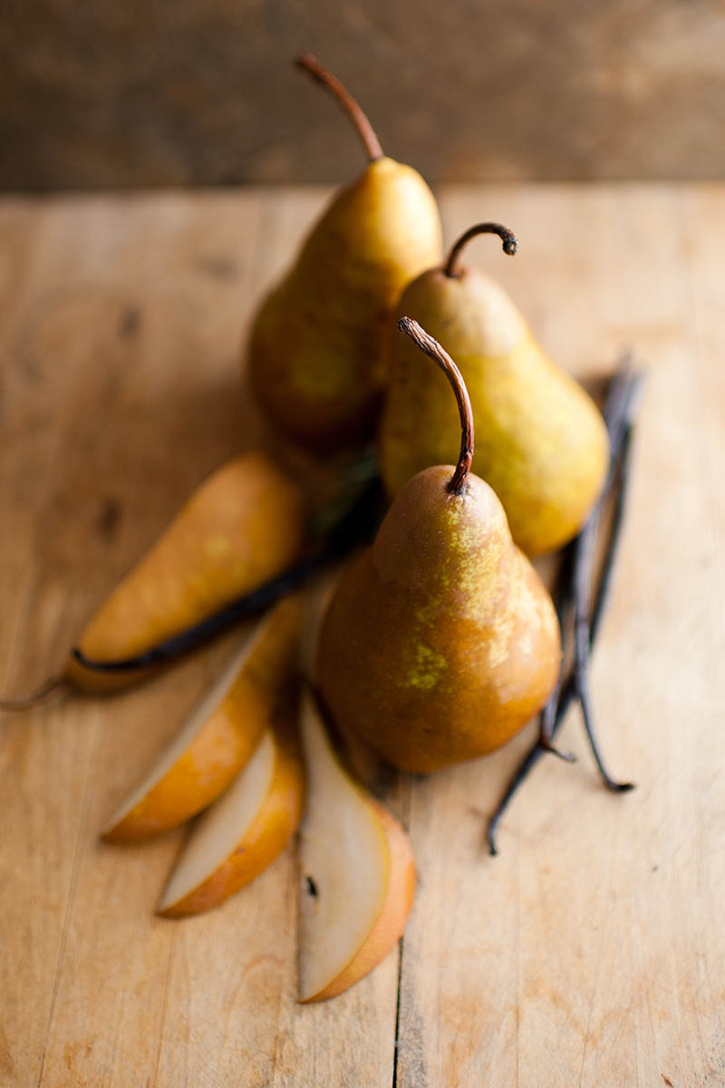 A close up vertical image of \'Bosc\' pears whole and sliced, set on a wooden chopping board with a few vanilla pods.